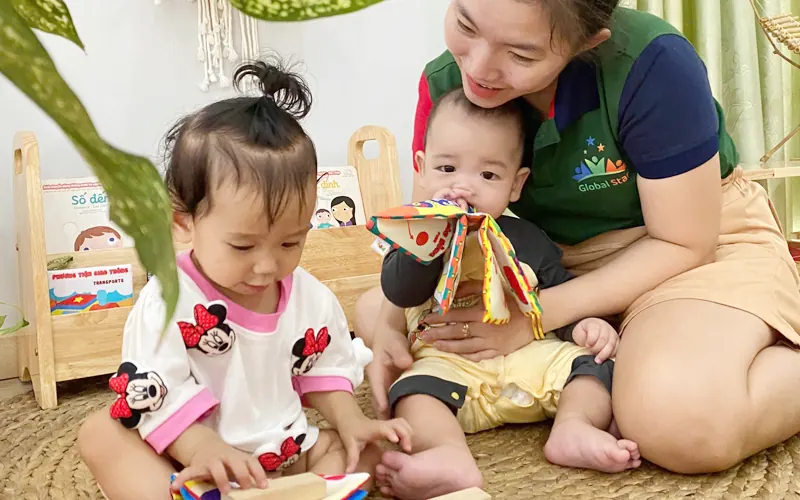 A teacher sits on a woven mat playing with two babies and a soft toy