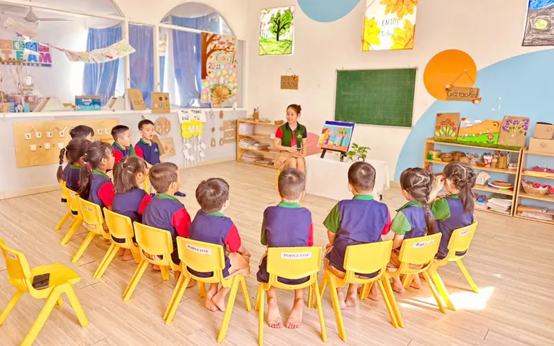A class sits on small yellow chairs in a circle around the teacher in a bright classroom